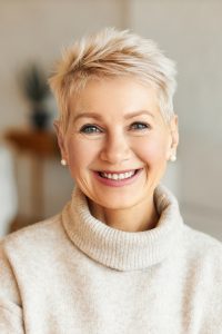 Close up image of happy good looking elegant fifty year old woman wearing warm cozy jumper, pearl earrings and short stylish hairdo being in good mood sitting in living room, smiling broadly at camera